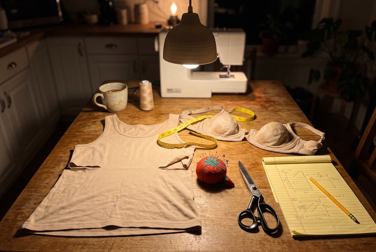 A kitchen table at night under a hanging lamp. A stretchy tank top cut apart at the seams, an old beige bra snipped in h